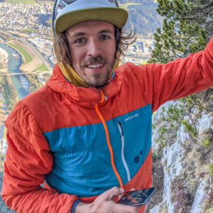 Kletterer in roter und blauer Jacke mit Helm an einer Felswand, dahinter Berglandschaft