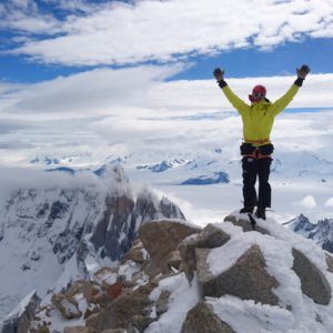 Person in gelber Jacke steht mit erhobenen Armen auf schneebedecktem Berggipfel mit Bergpanorama im Hintergrund