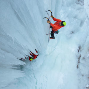 Eisklettererin in roter Jacke erklimmt steile gefrorene Wasserfallwand mit Eispickeln
