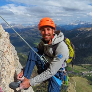 Kletterer in Sicherungsausrüstung an einer Felswand mit Bergpanorama im Hintergrund