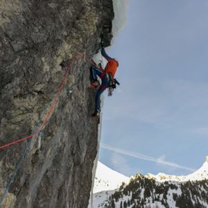 Kletterer in roter Ausrüstung an steiler Felswand mit verschneiten Bergen im Hintergrund