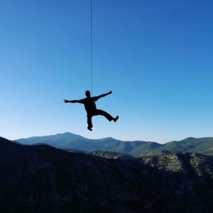 Silhouette einer Person springt an einem Seil über eine Berglandschaft bei blauem Himmel.